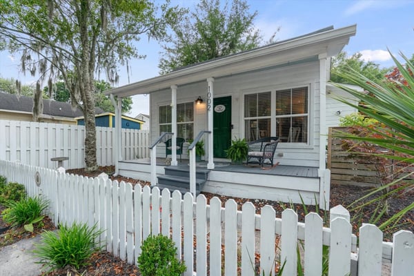 White cottage with green door and front porch, white picket fence, moss-draped oak trees, and tropical plants.