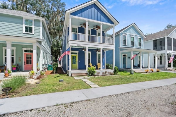 Row of modern cottage-style homes with blue and green siding, white porches, and American flags along a tree-lined street.