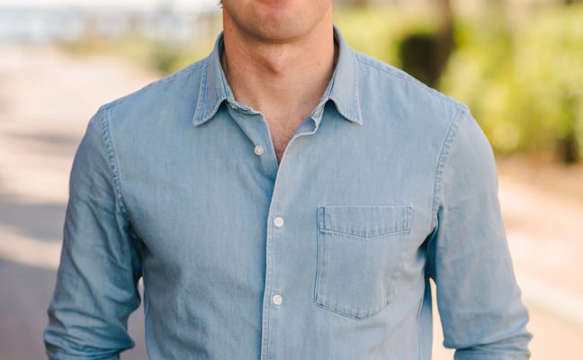 Man in light blue denim shirt standing outdoors with blurred trees and pathway in background