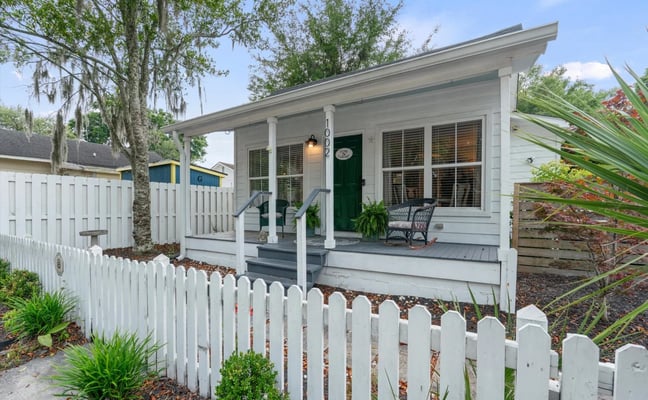 White cottage with green front door, covered porch, and white picket fence surrounded by oak trees and tropical plants